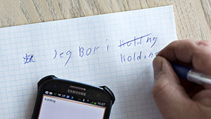 Hånd med pen skriver på papir med mobiltelefon ved siden af